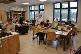 Pokaż zdjęcie: Pomieszczenie wielofunkcyjne. Widać skrawek kuchni i uczestniczkę, która maluje karton obok garnka. Przy dużym stole siedzą dziewiarki, które tworzą kolorowe makiety pod okiem instruktorki. Na trzecim stole warsztatowym leżą sznurki do makramy. P