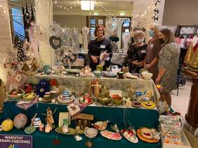 Pokaż zdjęcie: Fragment pomieszczenia w którym znajduje się bardzo dużo różnego rodzaju przedmiotów wykonanych z gliny, drewna, papieru oraz różnego rodzaju materiałów. Część przedmiotów wisi, a część jest poustawiana w rzędach na stołach. Obok stoją