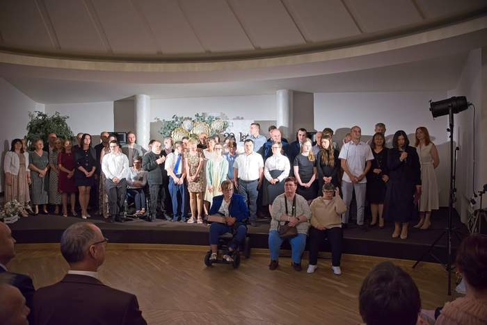 Pokaż zdjęcie: Grupa osób stoi na scenie podczas uroczystości w sali z kolumnami; z przodu siedzą uczestnicy, widoczna dekoracja kwiatowa i oświetlenie sceniczne, publiczność w tle.