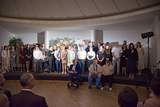 Grupa osób stoi na scenie podczas uroczystości w sali z kolumnami; z przodu siedzą uczestnicy, widoczna dekoracja kwiatowa i oświetlenie sceniczne, publiczność w tle.