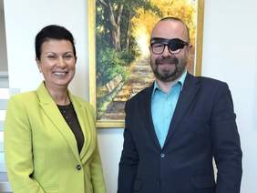 Pokaż zdjęcie: Kobieta i mężczyzna elegancko ubrani pozują do zdjęcia. W tle obraz z jesienną tematyką. mężczyzna ma czarną opaskę na jednym oku.