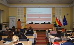 Pokaż zdjęcie: Zdjęcie przedstawia salę konferencyjną z uczestnikami siedzącymi przy stołach. Przy mównicy stoi kobieta wygłaszająca prelekcję, a za nią na ekranie wyświetlona jest prezentacja zatytułowana „Dostępna przestrzeń publiczna”. W tle znajdują się trzy flagi: Polski, województwa podlaskiego i Unii Europejskiej. Po lewej stronie widoczny jest baner Regionalnego Ośrodka Polityki Społecznej w Białymstoku. Przy stole prezydialnym siedzą trzy osoby, korzystające z laptopów.
