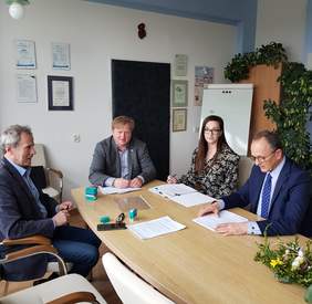 trzech mężczyzn i kobieta przy dużym jasnym stole na którym leżą dokumenty, jeden z mężczyzn i kobieta patrzą w obiektyw