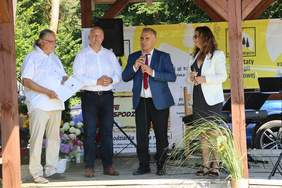 Pokaż zdjęcie: Trzech mężczyzn i kobieta stoją na scenie pod zadaszeniem. Jeden z nich ubrany w krawat biała koszule i marynarkę mówi do mikrofonu. Kobieta obok również trzyma mikrofon. W tle drzewa, słoneczna pogoda.