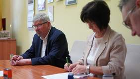 Pokaż zdjęcie: Przy stole siedzi kobieta i mężczyzna. Pani trzyma w rękach długopis i podpisuje dokument. Pan patrzy przed siebie. 