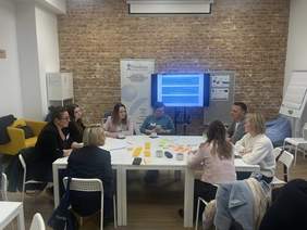 Pokaż zdjęcie: Grupa osób siedzących przy stole podczas warsztatów Fundacji Aktywizacja. Uczestnicy rozmawiają, piją kawę i pracują z karteczkami samoprzylepnymi. W tle ceglana ściana, baner fundacji i ekran z prezentacją.