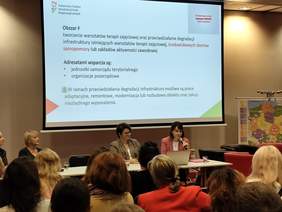 Pokaż zdjęcie: Sala wykładowa. Przy stole dla prelegentów siedzą kobiety, które prowadzą prelekcję. Na ekranie wyświetlone są informacje o programach PFRON. Widać tłum osób, który słucha wykładu i patrzy na slajdy. 