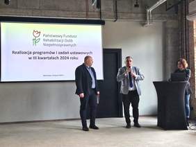 Pokaż zdjęcie: Na ekranie wyświetlana jest  prezentacja dotycząca realizacji programów PFRON. Dwóch mężczyzn przemawia, a kobieta obsługuje laptop. Wydarzenie odbywa się w nowoczesnym loftowym wnętrzu.