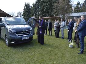 Pokaż zdjęcie: na posesji stoi samochód, który poświęcony jest przez księdza kropidłem obok stoją osoby i przyglądają się nowemu samochodowi