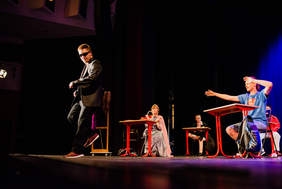 Pokaż zdjęcie: Chłopiec w garniturze i czarnych okularach tańczy na scenie. Obok siedzi chłopiec w niebieskiej koszulce Superman, za nimi dziewczynka ubrana w strój królewny