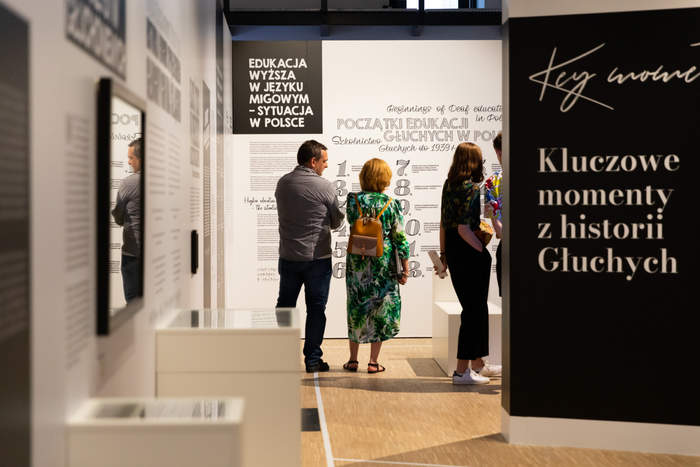 Pokaż zdjęcie: Fragment wysokiego pomieszczenia, na białe i czarne ściany są zamieszczone informacje o kluczowych momentach z historii Głuchych