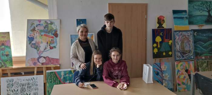 Pokaż zdjęcie: Dyrektor Oddziału w sali, w której znajdują się prace plastyczne, stoi obok młodzieży, która bierze udział w Grze Miejskiej