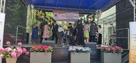 Scena plenerowa podczas festynu w Świdniku z okazji Dnia Godności Osób z Niepełnosprawnością Intelektualną. Na scenie grupa osób w strojach stylizowanych na dawne czasy, dekoracje kwiatowe i baner wydarzenia.