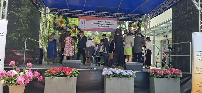 Pokaż zdjęcie: Scena plenerowa podczas festynu w Świdniku z okazji Dnia Godności Osób z Niepełnosprawnością Intelektualną. Na scenie grupa osób w strojach stylizowanych na dawne czasy, dekoracje kwiatowe i baner wydarzenia.