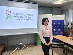 Pokaż zdjęcie: Na tle ekranu z logotypem Funduszu pozuje kobieta w różowej bluzce.