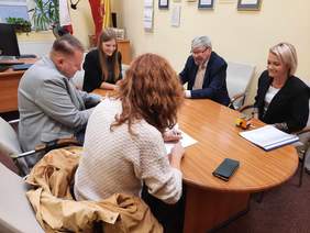 Pokaż zdjęcie: Przy stole siedzi pięć osób: trzy kobiety i dwóch mężczyzn. Podpisują umowę. Są uśmiechnięci. Na stole leżą dokumenty, pieczątki, telefon, długopis. 