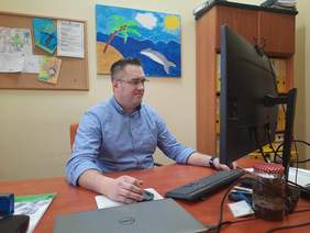 Pokaż zdjęcie: Przy biurku siedzi mężczyzna. Patrzy w monitor i delikatnie się uśmiecha. Dłonie trzyma obok klawiatury. Na ścianie wiszą kolorowe obrazki. 