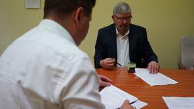 Pokaż zdjęcie: Przy stole siedzi dwóch panów. Jeden mężczyzna siedzi przodem do obiektywu aparatu, a drugi tyłem. Trzymają w rękach dokumenty. 