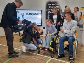 Pokaż zdjęcie: Dwie dziennikarki kucają przy dzieciach, z którymi przeprowadzają wywiad. Obok nich stoi operator, który nagrywa rozmowę kamerą. 