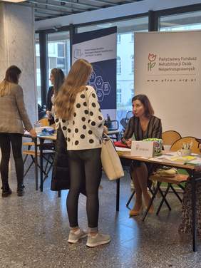 Pokaż zdjęcie: Na korytarzu uniwersytetu KUL stoją stoiska informacyjne. Przy stoliku siedzi kobieta, a przed nią stoi kobieta z długimi włosami i białym swetrze w czarne kółka. Kobiety rozmawiają.