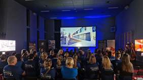 Pokaż zdjęcie: Grupa około sześćdziesięciu osób siedzi w sali konferencyjnej i ogląda na dużym ekranie premierę wirtualnego spaceru po SOSW w Kłecku. Po obu stronach ekranu widoczne są banery, w tym między innymi SOSW w Kłecku oraz Starostwa Powiatowego w Gnieźnie.