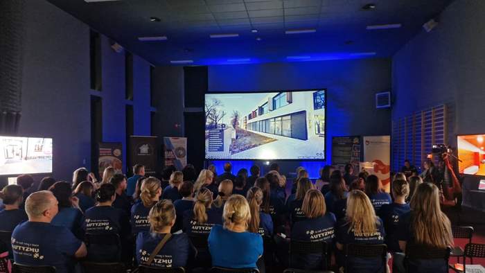 Pokaż zdjęcie: Grupa około sześćdziesięciu osób siedzi w sali konferencyjnej i ogląda na dużym ekranie premierę wirtualnego spaceru po SOSW w Kłecku. Po obu stronach ekranu widoczne są banery, w tym między innymi SOSW w Kłecku oraz Starostwa Powiatowego w Gnieźnie.