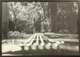 Pokaż zdjęcie: Zdjęcie wykonane z gitary, w centralnym punkcie zdjęcia widać rozmyte struny. W tle siedzi mężczyzna na wózku trzymający w uniesionym, prawym ręku gitarę.