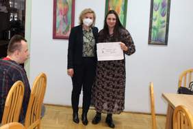 Pokaż zdjęcie: Dwie kobiety stoją w sali. Jedna z nich trzyma w dłoni białą torbę z czerwono- zielonym kwiatkiem i napisem Państwowy Fundusz Rehabilitacji Osób Niepełnosprawnych