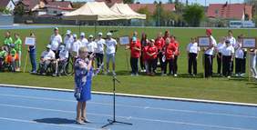 Pokaż zdjęcie: Kobieta trzyma w ręku mikrofon i przemawia. Za nią znajdują się drużyny sportowe. Wszyscy są na boisku