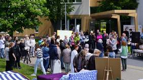 Pokaż zdjęcie: Przed budynkiem stoi kilkadziesiąt osób. Są to dzieci, kobiety, mężczyźni i osoby starsze. Jeden z mężczyzn przemawia do mikrofonu. 