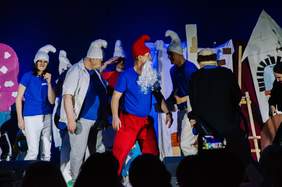 Pokaż zdjęcie: Na scenie występuje grupa teatralna. Aktorzy są przebrani za smerfy. Tańczą w kółku. Mają na sobie biało-niebieskie stroje i czapki smerfów. 