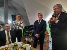 Pokaż zdjęcie: Przy wielkanocnym stole stoją trzy osoby: kobieta i dwóch mężczyzn. Jeden z panów przemawia do mikrofonu. Wszyscy się uśmiechają. W tle widać instrumenty muzyczne. 