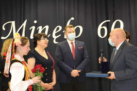 Pokaż zdjęcie: Mężczyzna trzyma mikrofon oraz medal Przyjaciela Szkoły. W tle mężczyzna oraz kobieta a także dzieci ubrane w strój krakowski trzymające czerwone róże 