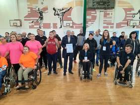 Pokaż zdjęcie: W hali sportowej znajduje się tłum ludzi. Część osób stoi, a inni siedzą na wózkach inwalidzkich. Wszyscy patrzą w stronę obiektywu aparatu. 