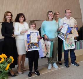 Pokaż zdjęcie: Sześć osób stoi razem podczas uroczystości wręczenia nagród. Troje uczestników trzyma dyplomy i kolorowe torby z upominkami, uśmiechając się do zdjęcia. Obok nich stoją trzy kobiety – przedstawicielki organizatorów i gości.