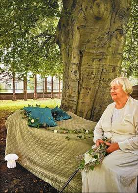 Pokaż zdjęcie: Fotografia przedstawiająca starszę kobietę w bieli siedzącą na ławce w parku i trzymającą kwiaty w dłoniach
