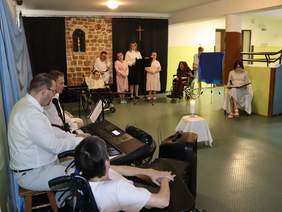 Pokaż zdjęcie: Grupa jedenastu osób, panowie po lewej stronie siedzą przy keabordzie, dwóch panów w przebraniu siedzi na wózkach inwalidzkich, cztery kobiety stoją przy mikrofonie i śpiewają, środek sali jest pusty.