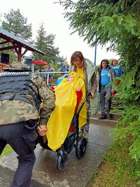 grupa ludzi, schodzą po szerokich schodach, kobieta i mężczyzna znosza osobę na wózku inwalidzkim, która ma żółta polerynę przeciwdeszczową