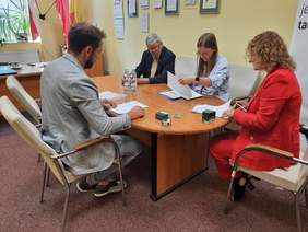 Pokaż zdjęcie: Przy owalnym stole siedzą cztery osoby: dwie kobiety i dwóch mężczyzn. Podpisują umowę. Na stole leżą różne dokumenty, pieczątki, woda mineralna. 