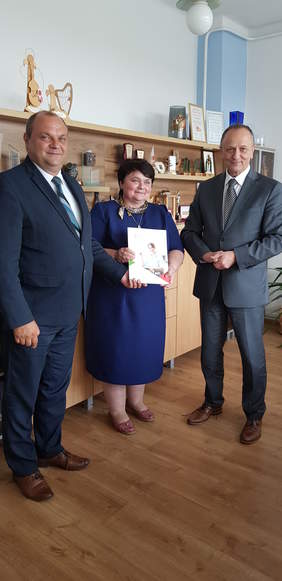 trzy osoby, od lewej: mężczyzna, kobieta, mężczyzna, stoją na tle mebli, uśmiechają się, kobieta trzyma w rękach teczkę z dokumentami