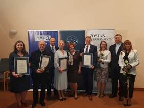 Zdjęcie grupowe na którym widzimy 9 osób, w tym 4 mężczyzn a wśród nich 2 księży, wszyscy elegancko ubrani, 4 osoby trzymają dyplomy Laureatów Konkursu Lodołamacze i statuetki, dwie panie trzymają po białej róży