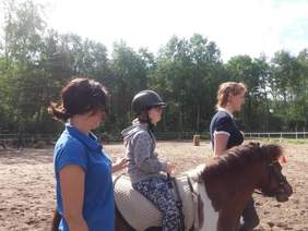 Pokaż zdjęcie: Dziecko jadące na kucyku prowadzonym przez terapeutkę. Dziecko jest podtrzymywane przez drugą osobę