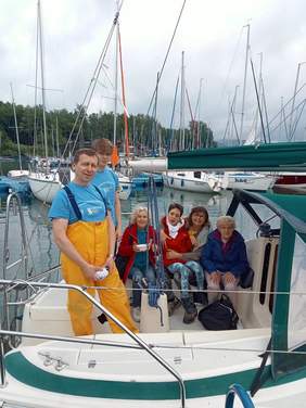 6 osób na łodzi, 2 mężczyżni w żółtych spodniach i niebieskich bluzkach stoją, 4 kobiety na prawo siedzą