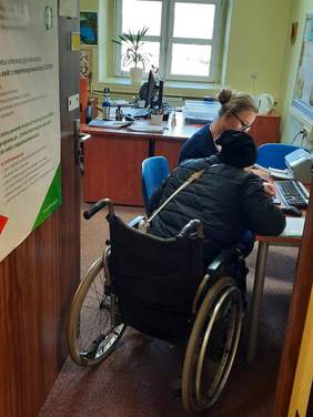 Pokaż zdjęcie: Pani na wózku inwalidzkim jest obsługiwana przez pracownicę. Po prawej stronie jest stanowisko komputerowe. Pracownik wprowadza dane do systemu. 