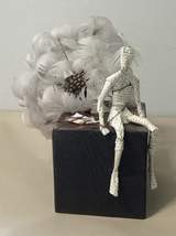 Papierowa figurka z tekstu siedząca na ciemnym drewnianym bloku, obok której znajduje się duża, kulista kompozycja z białych piór i drobnych elementów roślinnych.