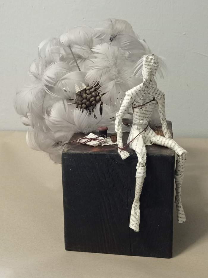 Pokaż zdjęcie: Papierowa figurka z tekstu siedząca na ciemnym drewnianym bloku, obok której znajduje się duża, kulista kompozycja z białych piór i drobnych elementów roślinnych.