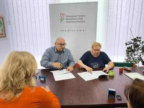 Pokaż zdjęcie: Zdjęcie dwóch osób siedzących przy stole podpisujących umowy, w tle rollup z logo Funduszu.