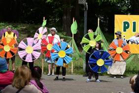 Pokaż zdjęcie: Grupa siedmiu osób ubranych w barwne stroje stoi na placu wyłożonym kostką brukową. Osoby te trzymają w dłoniach różnokolorowe obręcze, do których to przymocowany został materiał w kolorze danej obręczy, imitujący duży kwiat.