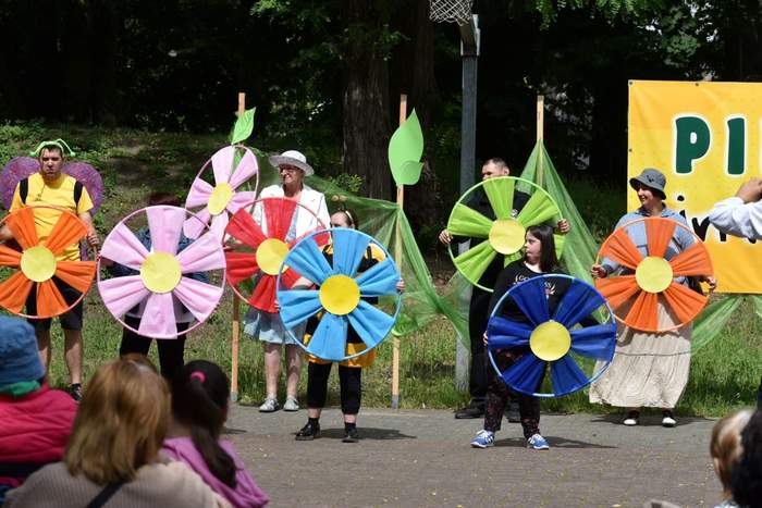 Pokaż zdjęcie: Grupa siedmiu osób ubranych w barwne stroje stoi na placu wyłożonym kostką brukową. Osoby te trzymają w dłoniach różnokolorowe obręcze, do których to przymocowany został materiał w kolorze danej obręczy, imitujący duży kwiat.