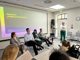 Pokaż zdjęcie: Prelegenci przed publicznością w sali konferencyjnej z wyświetlonym tytułem warsztatów "Prawo do Dostępności" na ekranie.